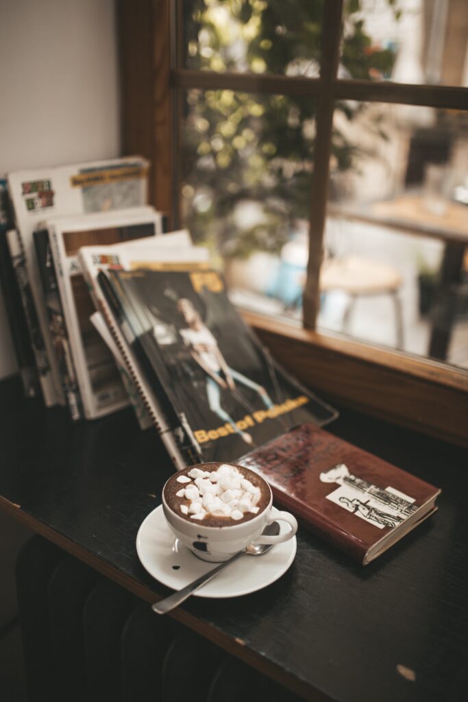 Image of a white ceramic teacup on a saucer full of hot chocolate and marshmallows, with out of focus books and a window behind it.